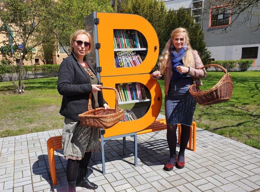 Zdjęcie bibliotecznego skweru, na krórym stoi półka bookcrossingowa w kształcie litery B. Wokół placyku wyłożonym kostką polbruk widac zieleń miejską. Dwie bibliotekarki stoją obok półki