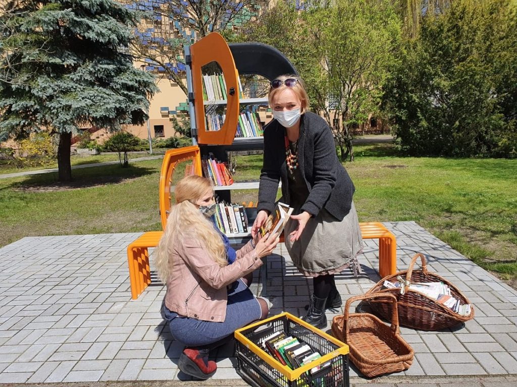 Zdjęcie bibliotecznego skweru, na krórym stoi półka bookcrossingowa w kształcie litery B. Wokół placyku wyłożonym kostką polbruk widać zieleń miejską. Dwie bibliotekarki układają w niej książki
