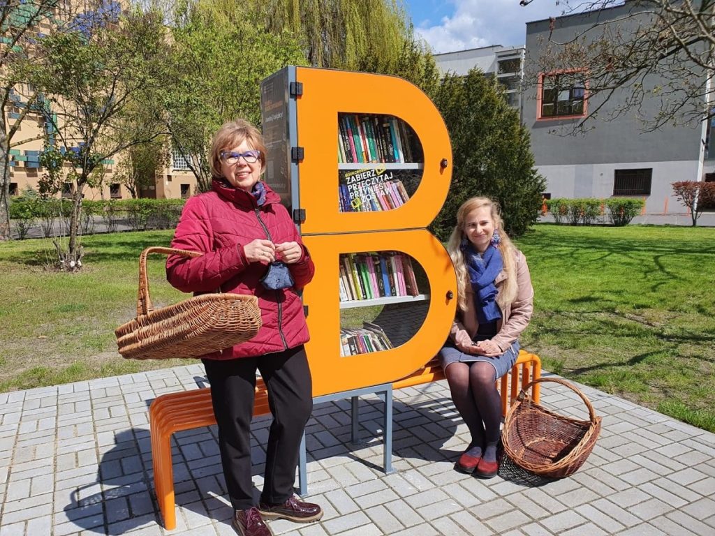 Zdjęcie bibliotecznego skweru, na krórym stoi półka bookcrossingowa w kształcie litery B. Wokół placyku wyłożonym kostką polbruk widac zieleń miejską. Obok półki, po lewej stronie stoi dyrektor MiP Biblioteki Publicznej w Nowym Tomyślu, po prawej siedzi jedna z bibliotekarek