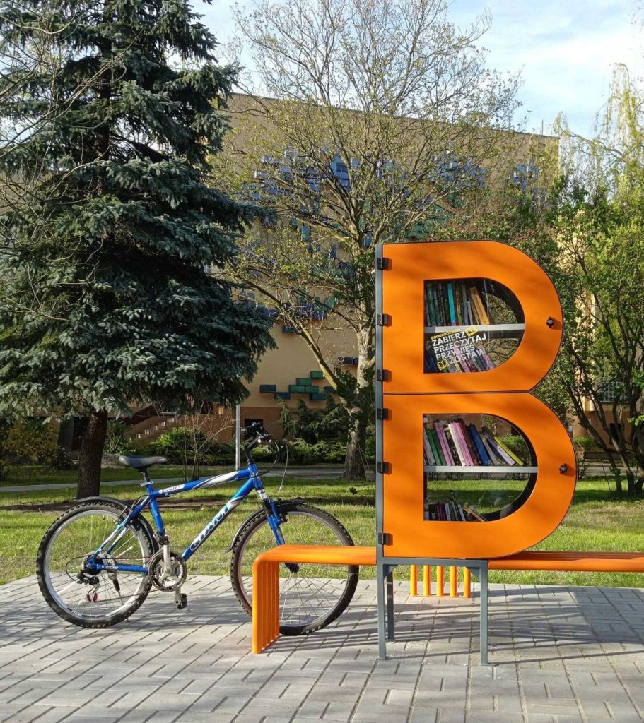 Zdjęcie bibliotecznego skweru, na króeym stoi półka bookcrossingowa w kształcie litery B. Wokół placyku wyłożonym kostką polbruk. Przy regale, po lewej stronie stoi rower