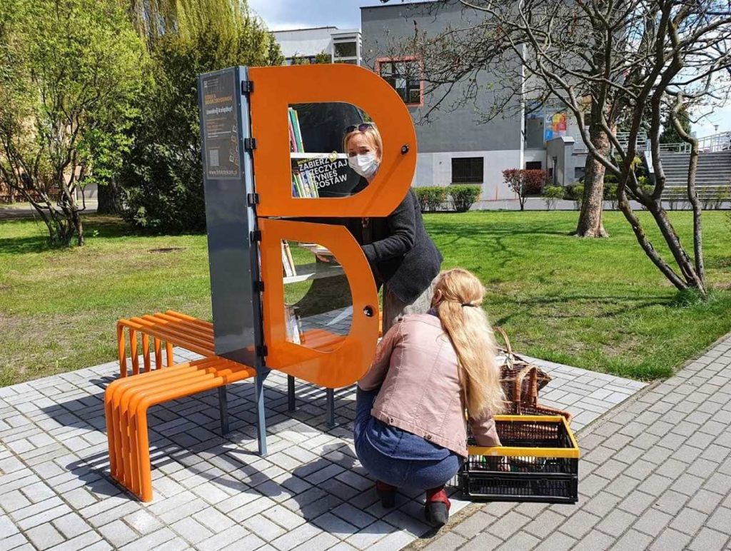 Zdjęcie bibliotecznego skweru, na krórym stoi półka bookcrossingowa w kształcie litery B. Wokół placyku wyłożonym kostką polbruk widać zieleń miejską. Dwie bibliotekarki układają w niej książki