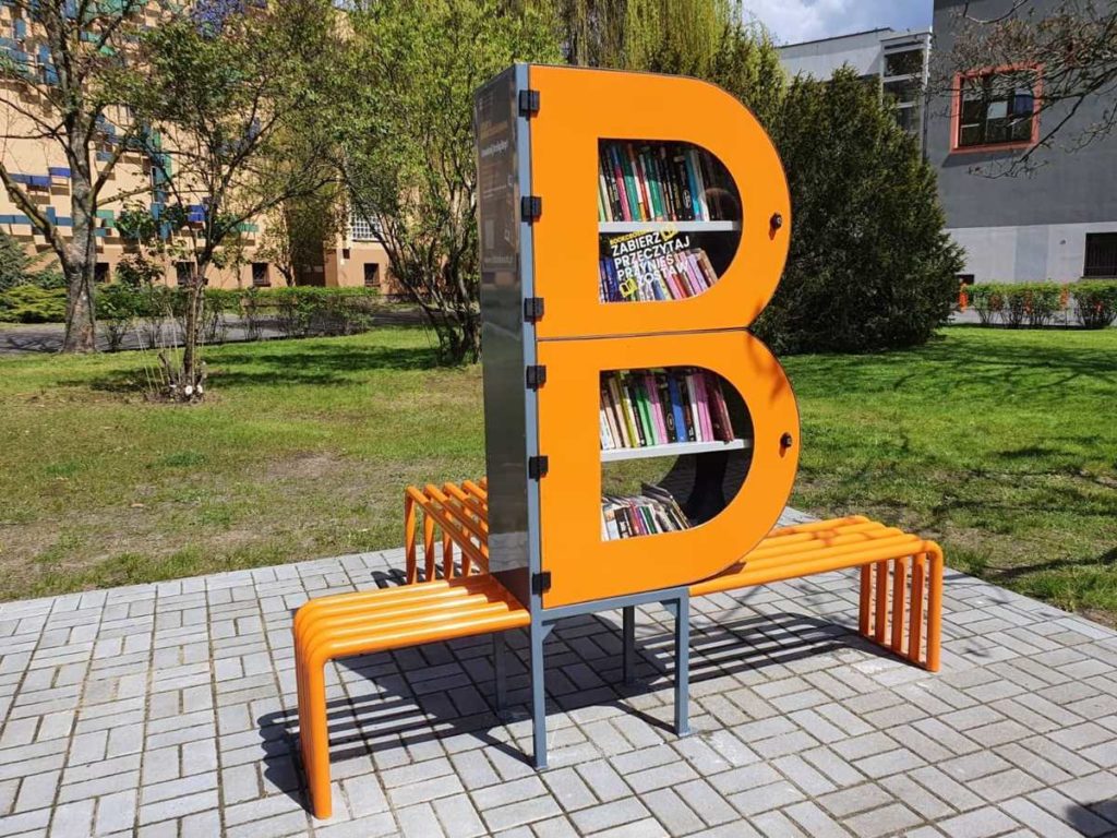 Zdjęcie bibliotecznego skweru, na króeym stoi półka bookcrossingowa w kształcie litery B. Wokół placyku wyłożonym kostką polbruk widac zieleń miejską