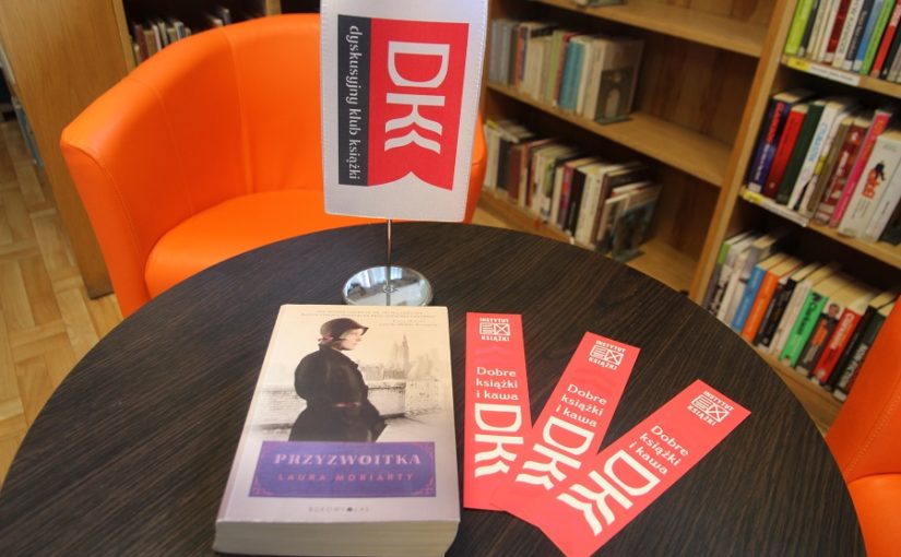 na pierwszym planie, na stoliku, książka i zakładki klubu DKK, w tle regał z książkami i pomarańczowy fotel