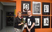 zdjęcie przedstawia dwie uśmiechnięte osoby w okularach, w tle ściana na której wiszą portrety czarno-białe.